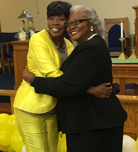 two women hugging, one in yellow and the other in black, in front of balloons